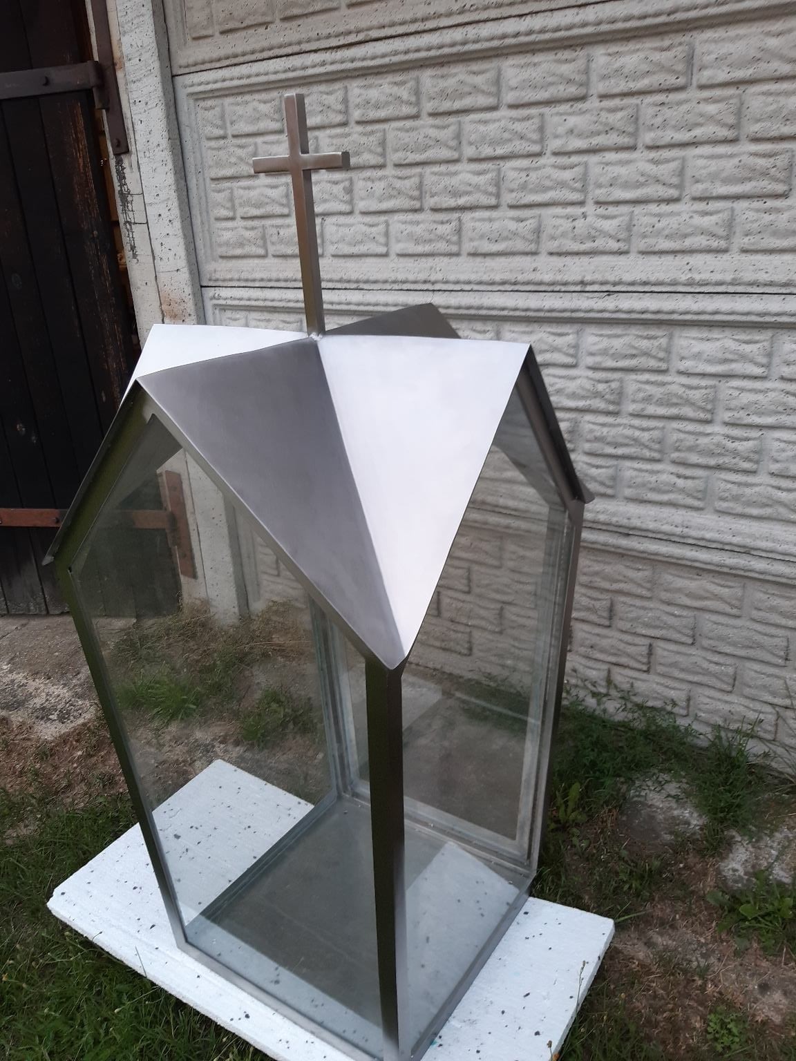Nierdzewna kapliczka z krzyżem na szczycie, szklane ścianki, tło z ceglanego muru i drewnianych drzwi.