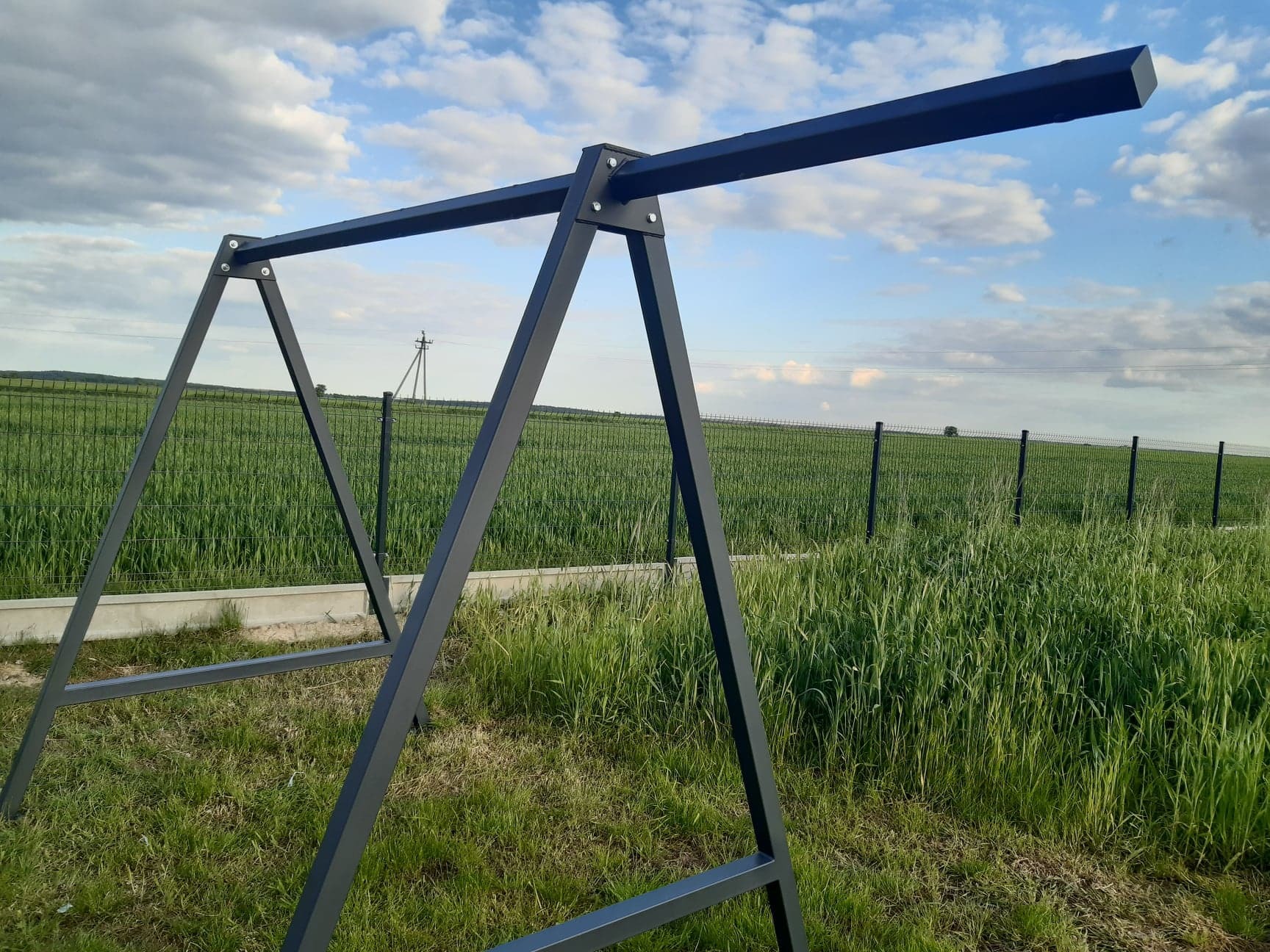 Metalowa konstrukcja huśtawki ogrodowej w kolorze grafitowym, ustawiona na trawniku z widokiem na pole uprawne i ogrodzenie panelowe w tle.