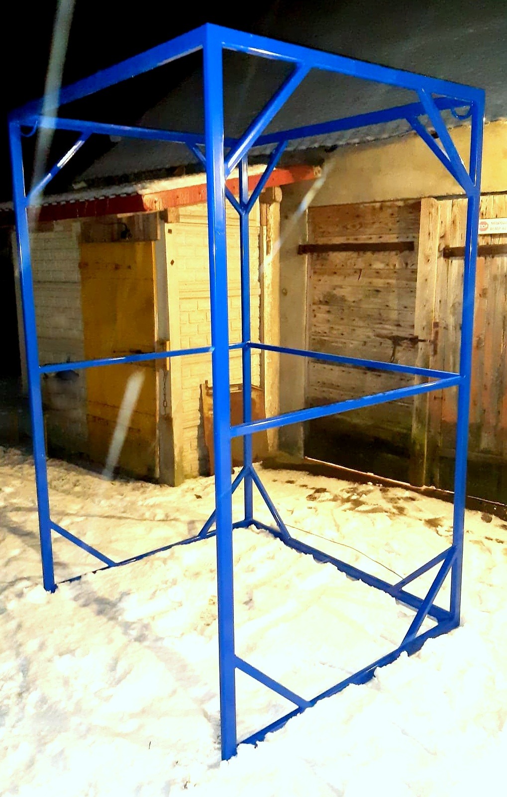 Niebieska, metalowa konstrukcja szkieletowa na śniegu, przypominająca zarys altany lub pergoli, z drewnianymi budynkami w tle.