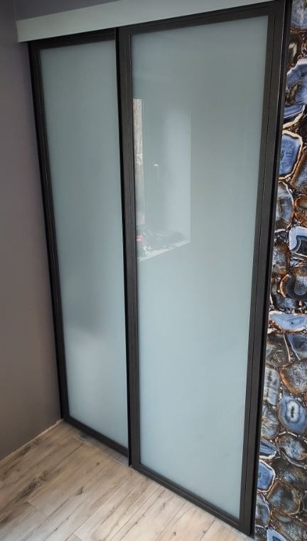 Dwie przesuwne szklane drzwi z ciemną ramą, jedna z nich częściowo zasłania zawartość szafy, obok ściana wykończona płytkami imitującymi przekrój agatu.