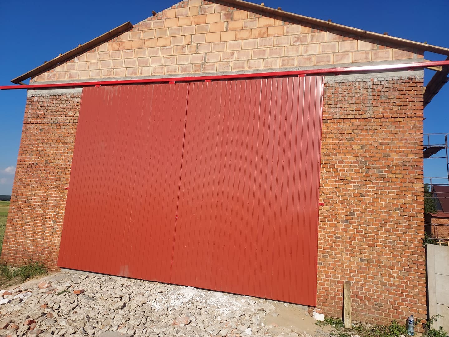 Duża, czerwona, przesuwna brama z blachy falistej zamontowana w ceglanym budynku w trakcie budowy, widoczne rusztowania i gruz.