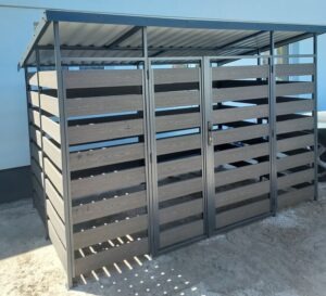 Zadaszona konstrukcja metalowa z wypełnieniem z paneli kompozytowych imitujących drewno, tworząca osłonę na śmietniki lub inne urządzenia, widoczna z boku, na betonowym podłożu.