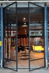 Otwarta, dwuskrzydłowa brama z czarną, metalową ramą i przezroczystym wypełnieniem szklanym, prowadząca do wnętrza budynku. Widoczne elementy konstrukcyjne i wyposażenie wnętrza w tle.