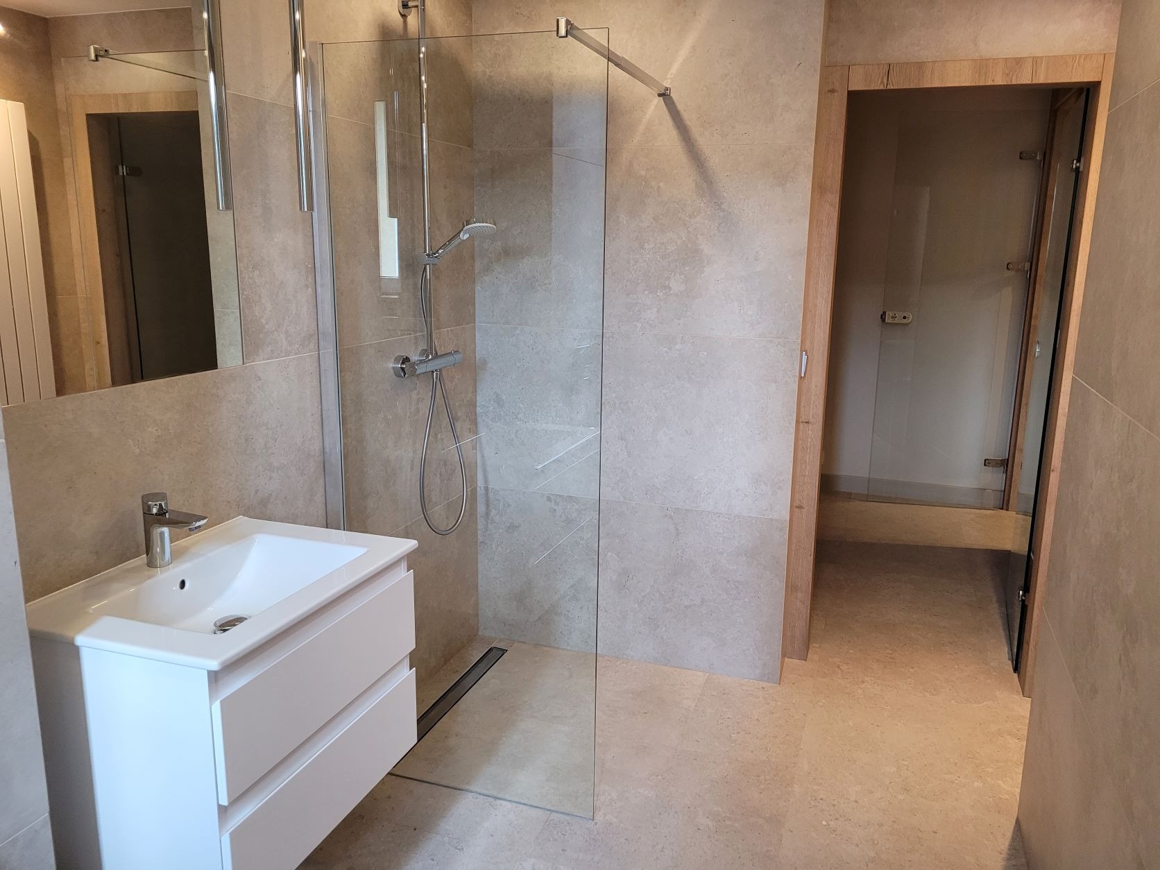 Nowoczesna łazienka z beżowymi płytkami, białą szafką pod umywalką, szklaną kabiną prysznicową bez brodzika i drewnianą ramą drzwi.