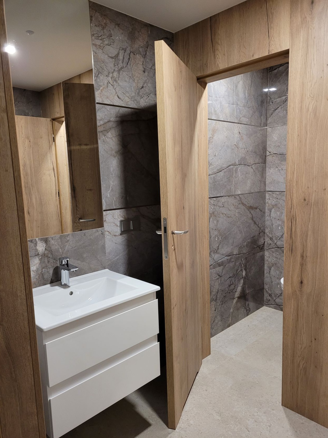 Nowoczesna łazienka z umywalką nablatową na białej szafce, marmurowymi płytkami na ścianach i drewnopodobnymi drzwiami oraz obudową.
