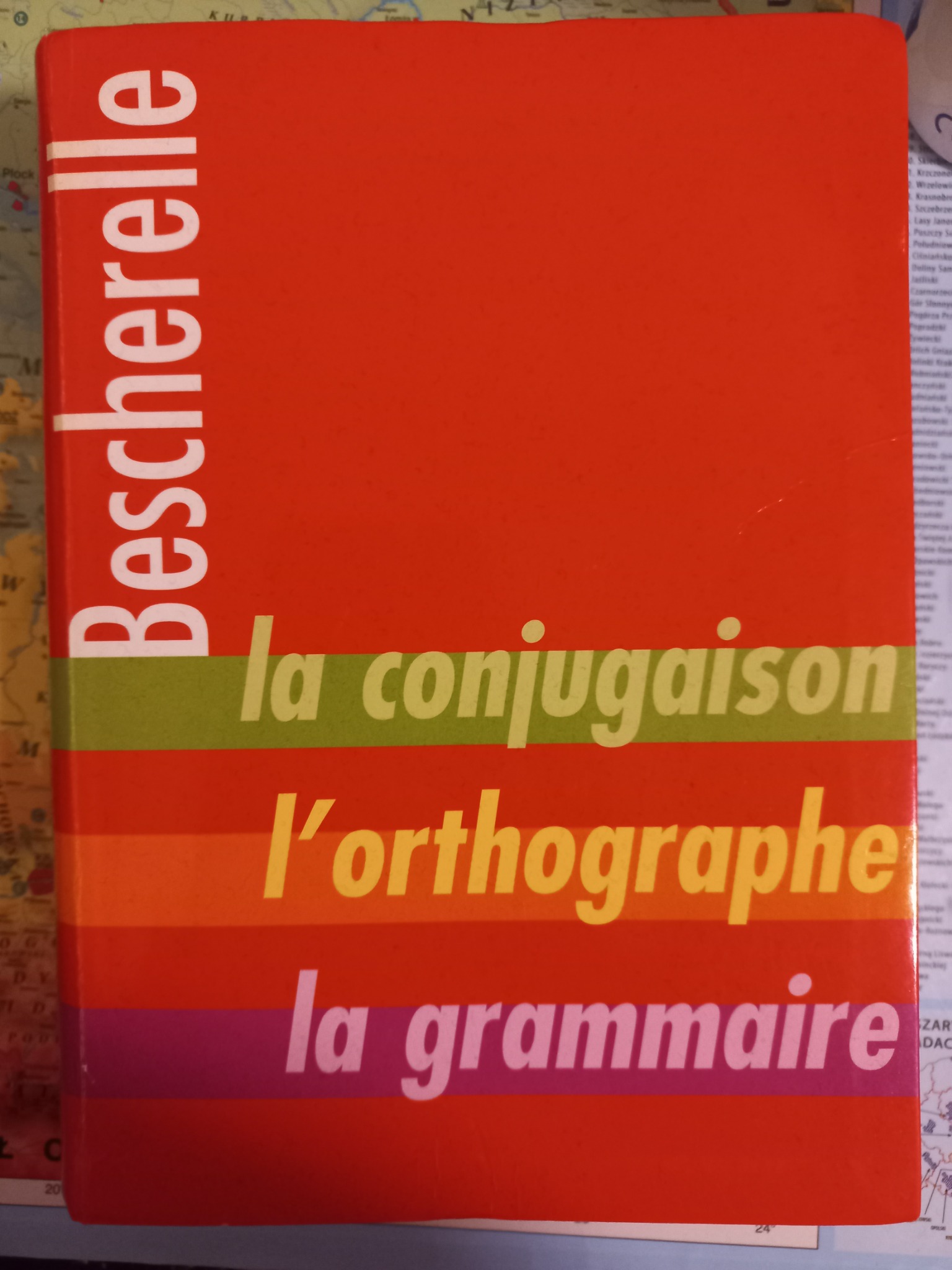 Czerwony podręcznik do nauki języka francuskiego Bescherelle, z tytułami: la conjugaison, l'orthographe, la grammaire, leżący na mapie.