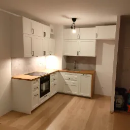 Biała, narożna zabudowa kuchenna z drewnianymi blatami, czarnymi uchwytami i płytą indukcyjną, oświetlona punktowo, na parkiecie.