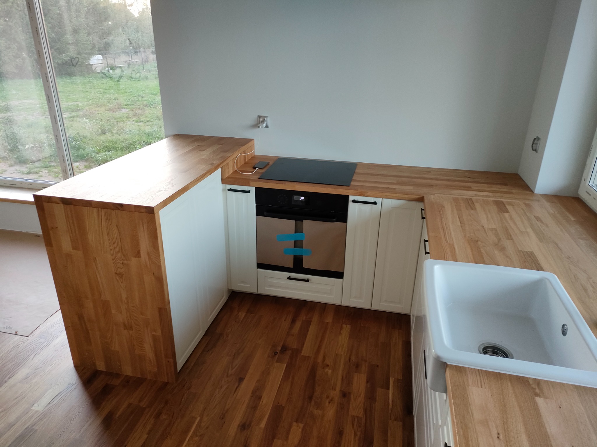 Nowoczesna kuchnia z białymi szafkami i drewnianym blatem, widok na okno z ogrodem w tle, na blacie płyta indukcyjna i ładowarka do telefonu.