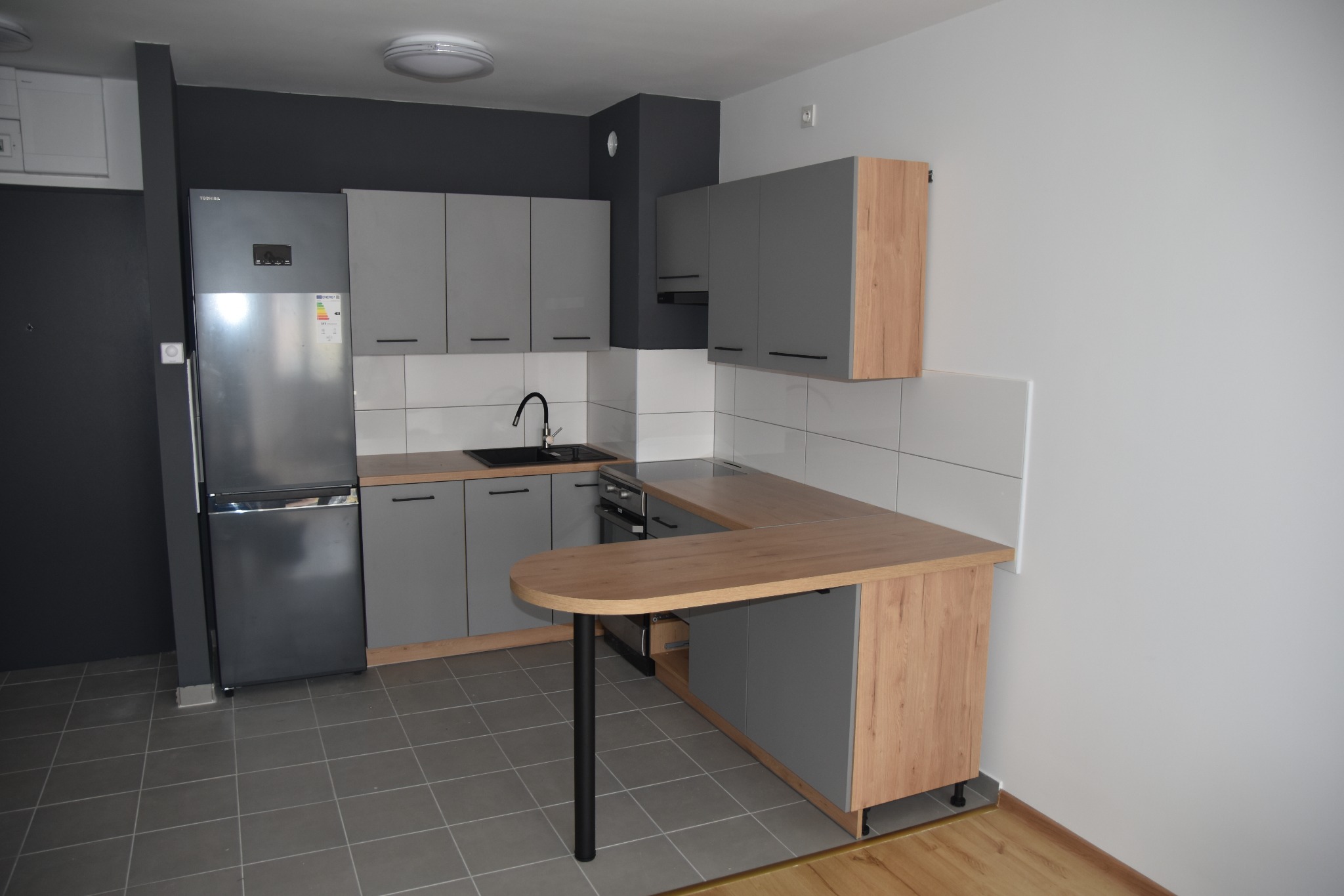 Nowoczesna kuchnia w szarościach z drewnianym blatem i półwyspem, widok na zabudowę kuchenną, płytki na ścianie i szarą podłogę.