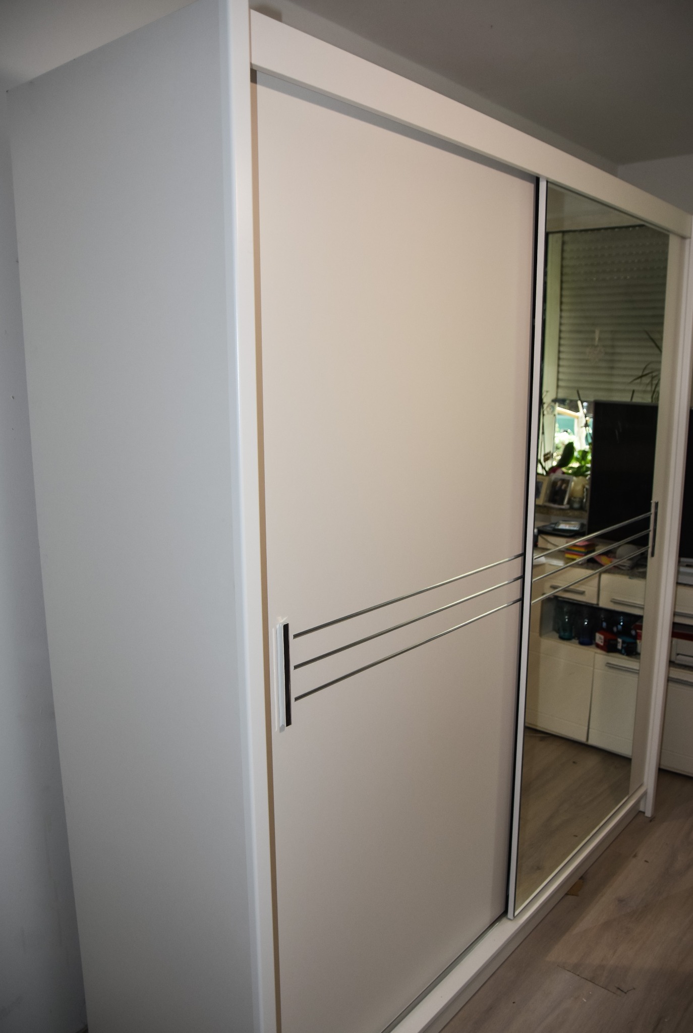 Biała szafa przesuwna z lustrem, widoczne trzy poziome, chromowane uchwyty na drzwiach, odbicie pokoju w lustrze.