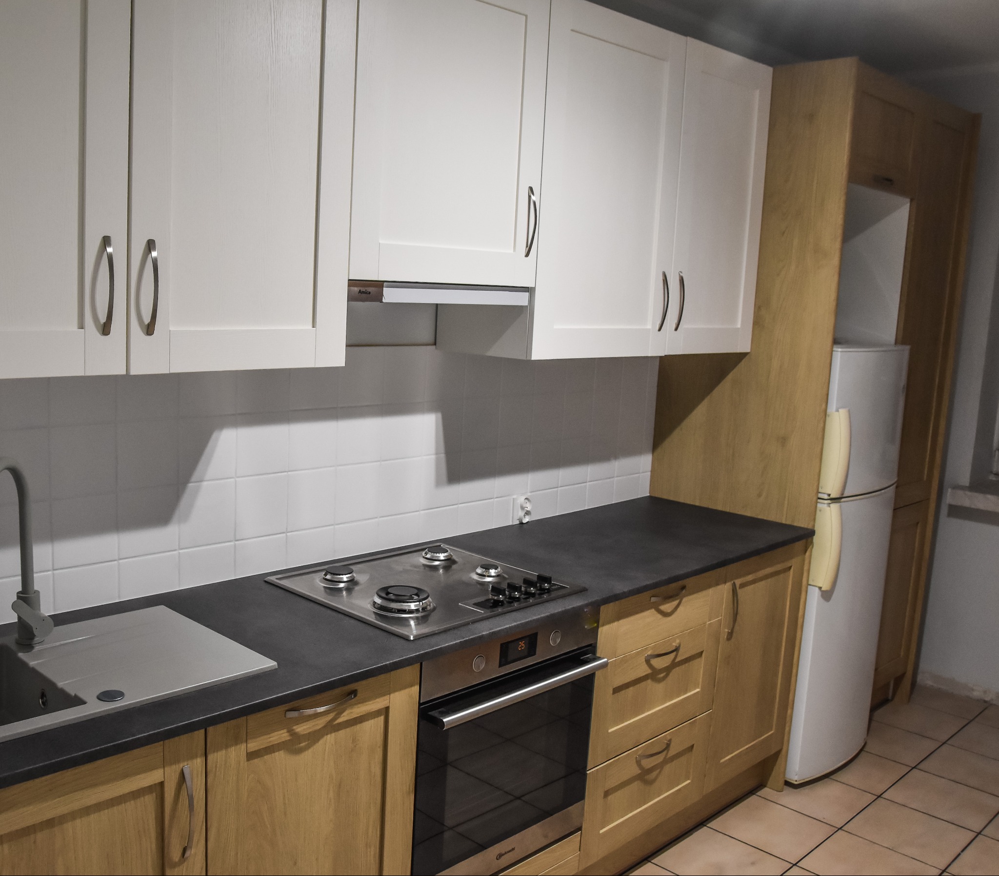 Jasna kuchnia z białymi szafkami górnymi i drewnianymi dolnymi, czarny blat, płyta gazowa i piekarnik ze stali nierdzewnej, białe kafelki na ścianie.