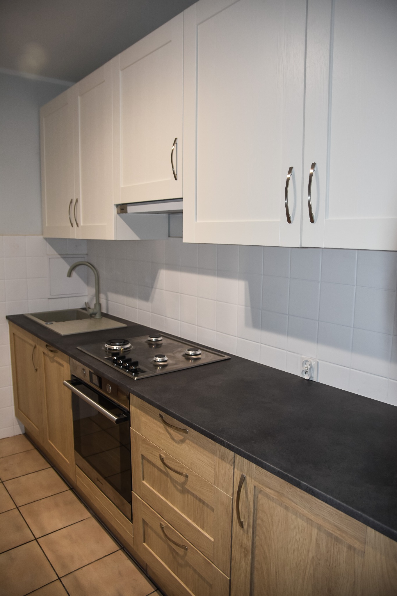 Nowoczesna kuchnia z białymi szafkami wiszącymi i drewnianymi szafkami dolnymi, czarny blat z wbudowaną płytą grzewczą, widoczny zlew i bateria, białe kafelki na ścianie.