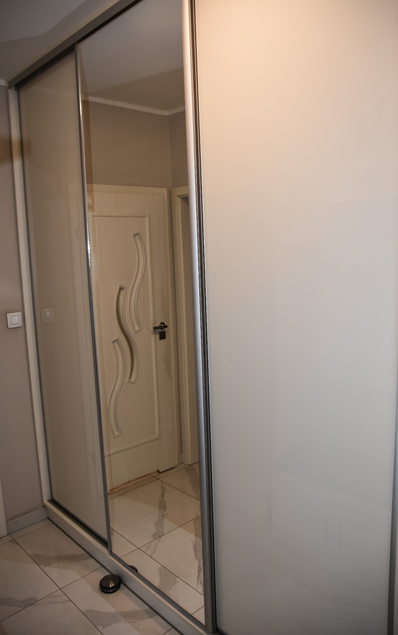 Biała szafa przesuwna z lustrem, odbijająca drzwi z dekoracyjnym wzorem, na marmurowej podłodze.