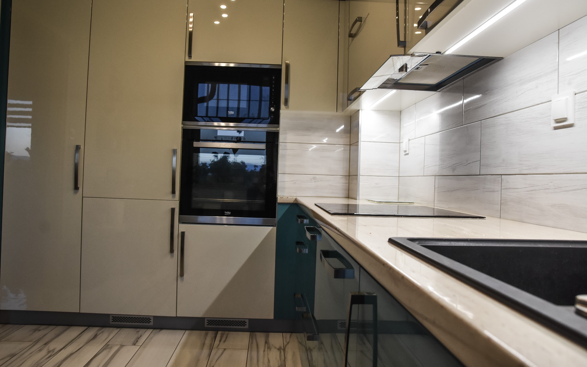 Nowoczesna kuchnia na wymiar z beżowymi, lakierowanymi frontami, zabudowanym piekarnikiem i mikrofalówką marki Beko oraz szklaną płytą indukcyjną, oświetlona listwą LED pod szafkami wiszącymi.