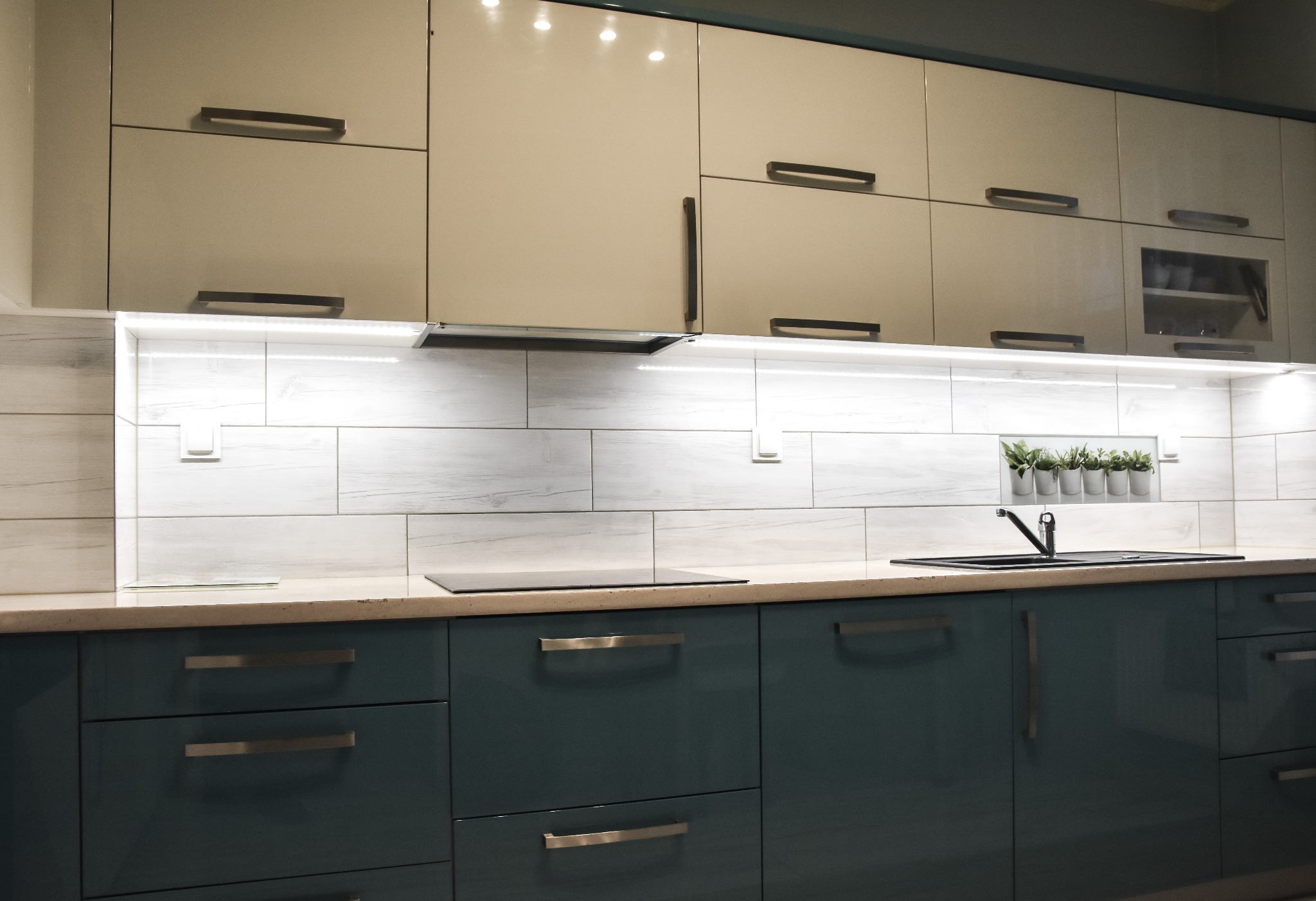 Nowoczesna kuchnia z górnymi szafkami w odcieniach beżu i dolnymi w kolorze ciemnej zieleni. Białe płytki imitujące drewno na ścianie nad blatem, podświetlenie LED i czarny zlew.