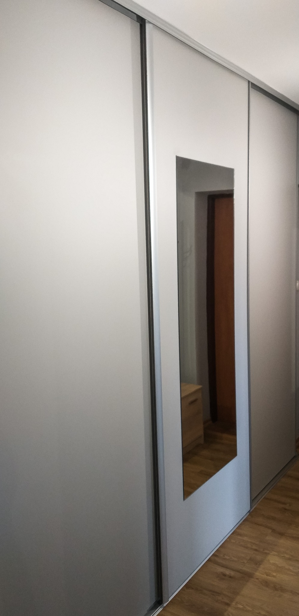 Szara szafa przesuwna z lustrem wbudowanym w jedno ze skrzydeł, odbijającym fragment wnętrza pokoju z drewnianą podłogą i szafką.
