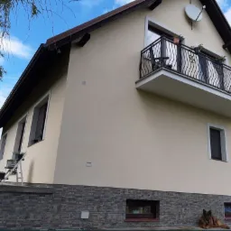 Elewacja domu jednorodzinnego po remoncie, z beżową ścianą, balkonem z kutą balustradą i kamienną podmurówką, na której leży pies.