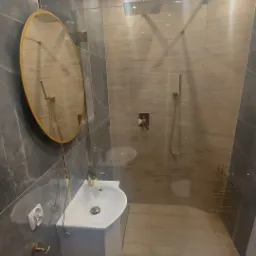 Nowoczesna łazienka z szarymi płytkami imitującymi marmur, kabiną prysznicową typu walk-in, owalnym lustrem w złotej ramie i białą umywalką z szafką, wykończona złotymi detalami.