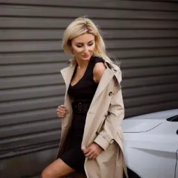 Blondwłosa kobieta w beżowym trenczu i czarnej sukience pozuje oparta o biały samochód na tle szarej ściany.