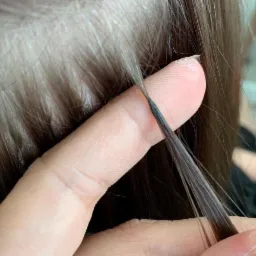 Doczepianie ciemnych pasemek do brązowych włosów, widoczne łączenie przy skórze głowy, palec przytrzymuje pasemko.