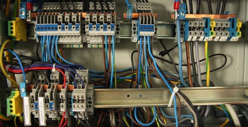 Rozdzielnica elektryczna z widocznymi bezpiecznikami, złączkami i przewodami w różnych kolorach, zamocowana na szynie DIN.