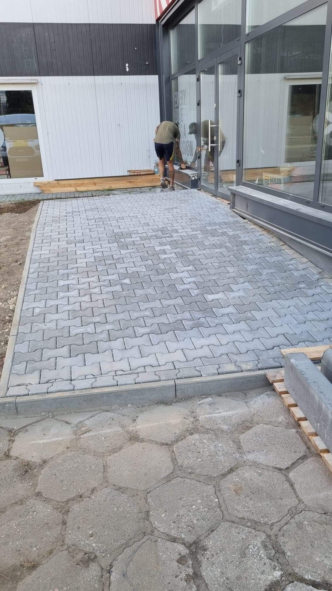 Pracownik układa szare kostki brukowe typu 'kość' na chodniku przed budynkiem z przeszklonym frontem; widoczne obrzeża chodnika, fragment starej nawierzchni z sześciokątnych płyt oraz paleta...