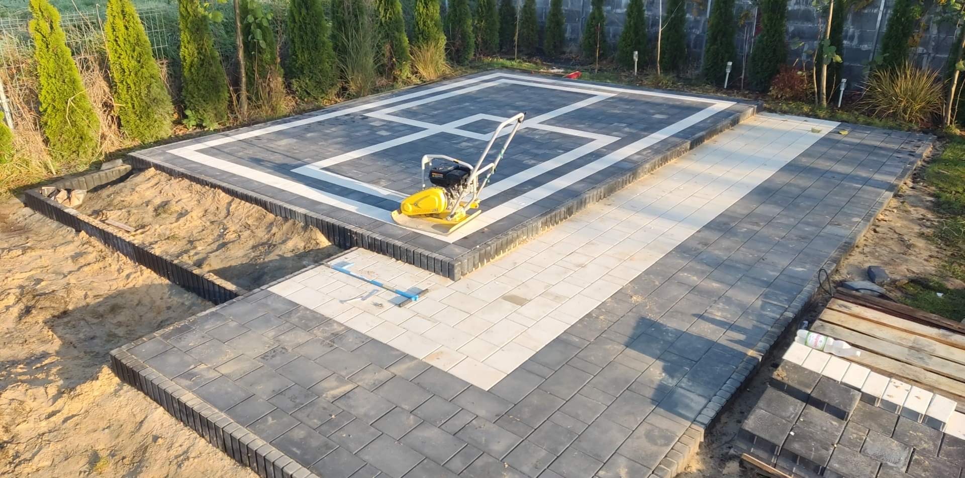 Widok z góry na częściowo ułożony taras z kostki brukowej w odcieniach szarości i bieli, z zagęszczarką w centralnej części i przygotowanym wykopem obok, otoczony zielenią.