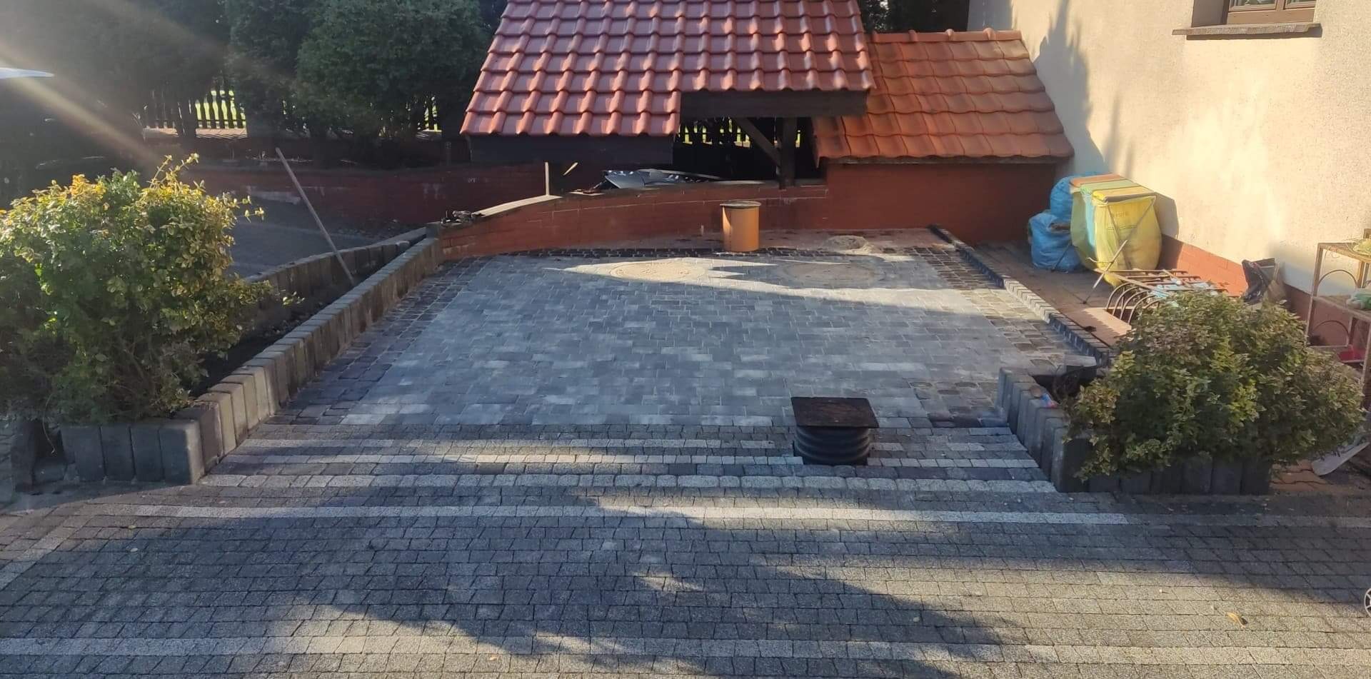 Ułożona szara kostka brukowa na podjeździe z niskimi schodami, obrzeża z betonowych elementów, w tle zadaszenie z czerwoną dachówką i fragment budynku.