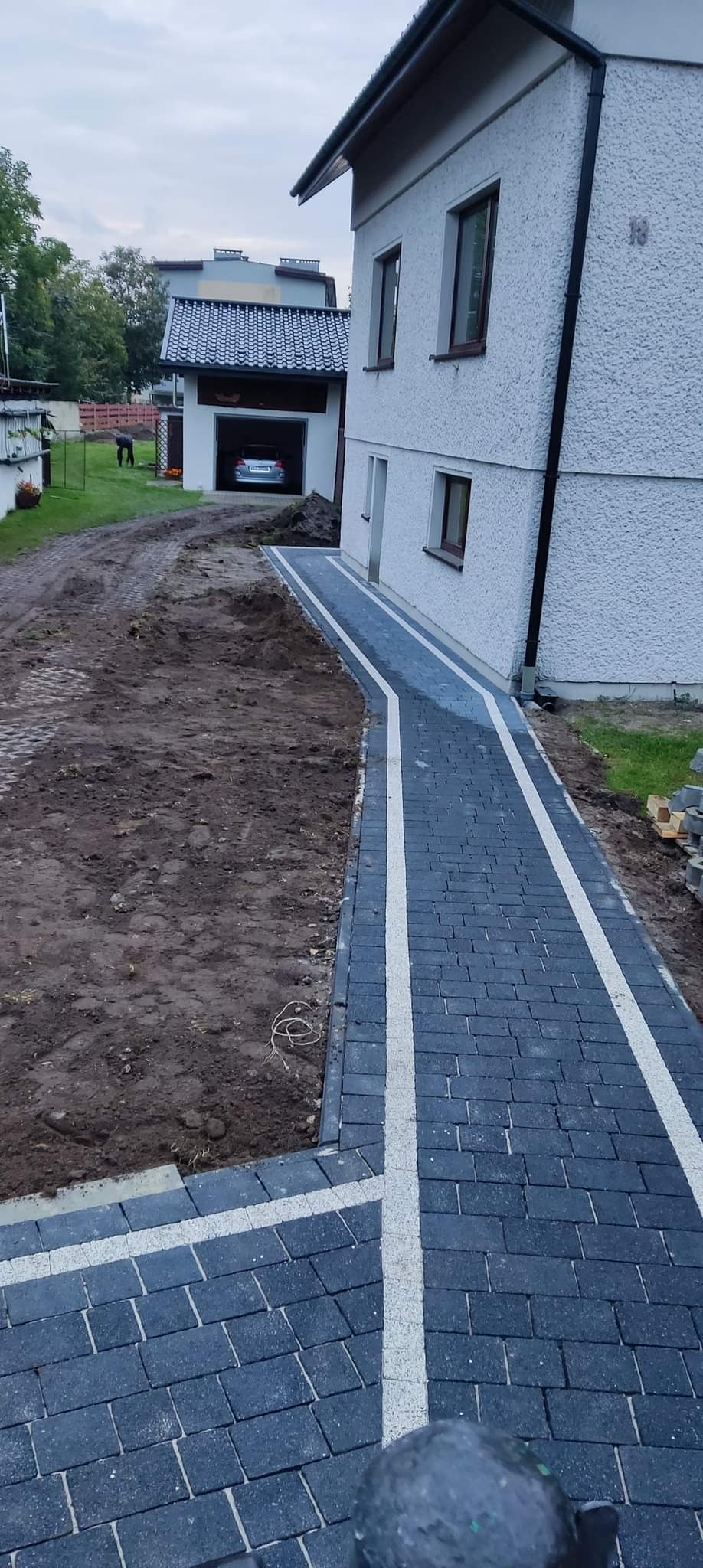 Ułożona ścieżka z ciemnej kostki brukowej z białymi akcentami prowadząca do budynku z garażem, widoczne przygotowane podłoże gruntowe po lewej stronie, częściowo ułożone kostki po prawej stronie.