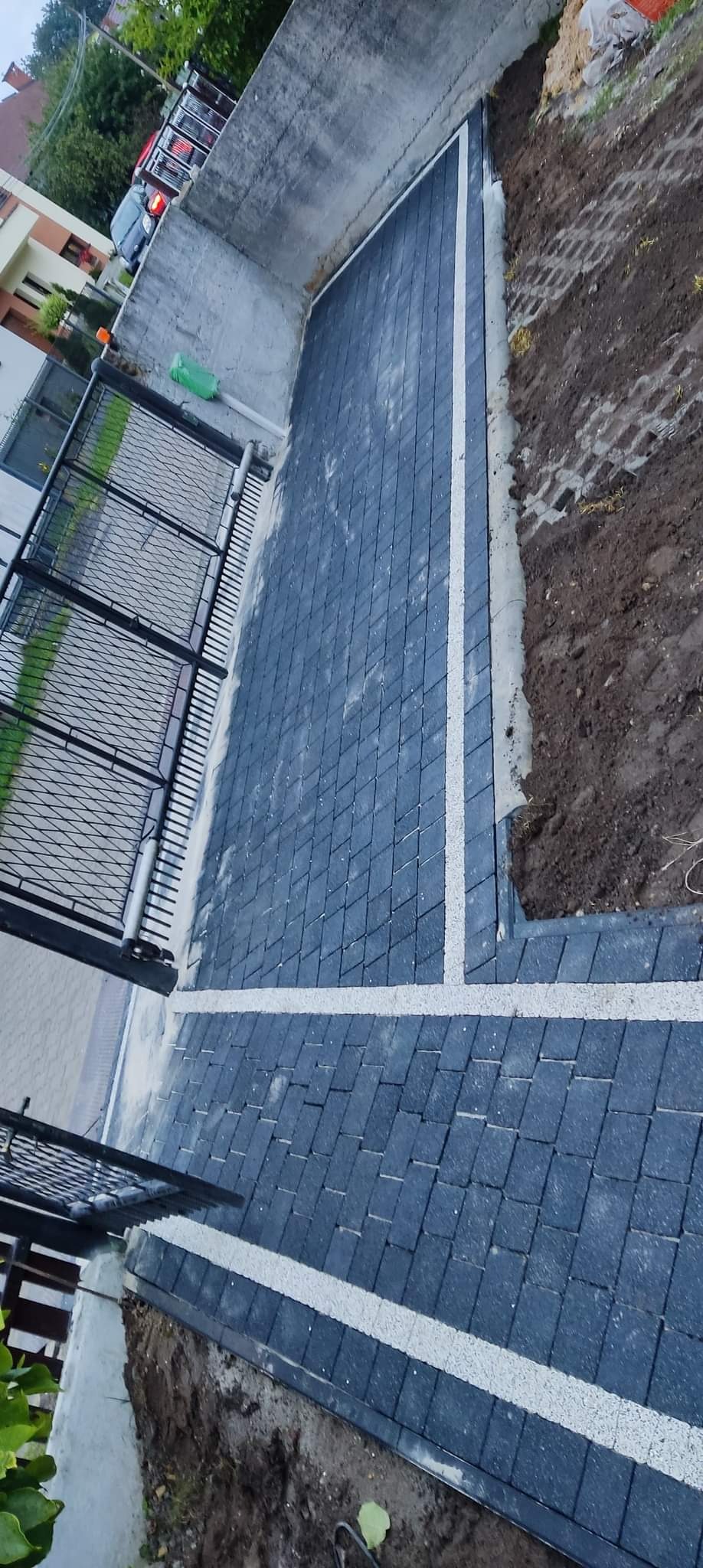 Niedawno ułożona, grafitowa kostka brukowa z jasnymi pasami oddzielającymi, widoczna brama wjazdowa i świeżo obsypana ziemia.
