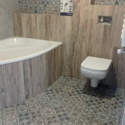 Wykończona łazienka z narożną wanną, wiszącą toaletą i dekoracyjnymi płytkami podłogowymi w stylu vintage, drewnopodobne panele ścienne, pudełko z deszczownicą na ścianie.