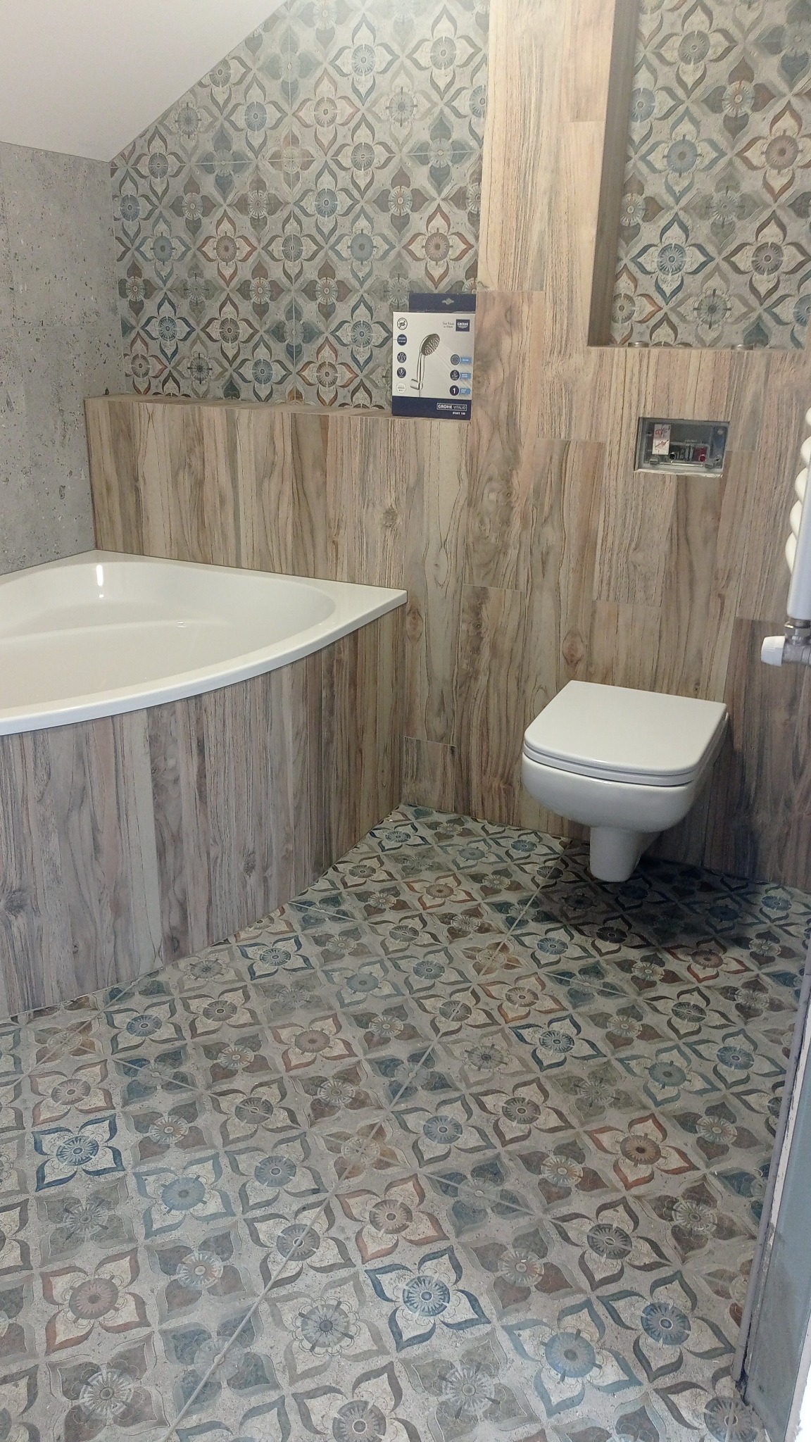 Wykończona łazienka z narożną wanną, wiszącą toaletą i dekoracyjnymi płytkami podłogowymi w stylu vintage, drewnopodobne panele ścienne, pudełko z deszczownicą na ścianie.