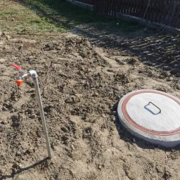Zakończona instalacja: metalowa rura z zaworem i pomarańczową końcówką wystaje z ziemi obok betonowej pokrywy studni z uchwytem, otoczone świeżo przekopaną ziemią i śladami opon.