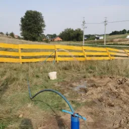 Woda tryskająca z niebieskiej rury wystającej z ziemi, z żółtym ogrodzeniem w tle, podczas wiercenia studni na zewnątrz.