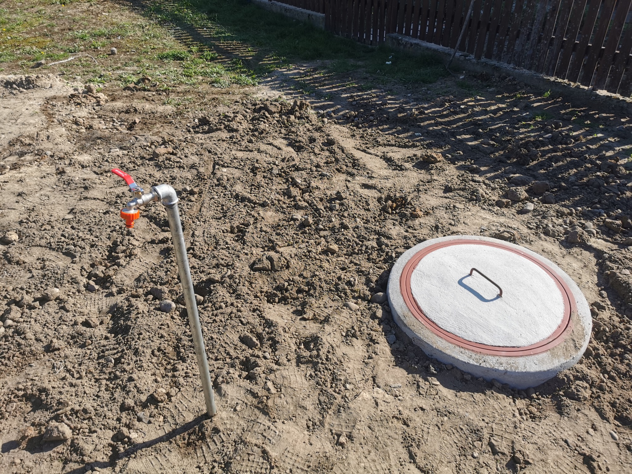 Zakończona instalacja: metalowa rura z zaworem i pomarańczową końcówką wystaje z ziemi obok betonowej pokrywy studni z uchwytem, otoczone świeżo przekopaną ziemią i śladami opon.