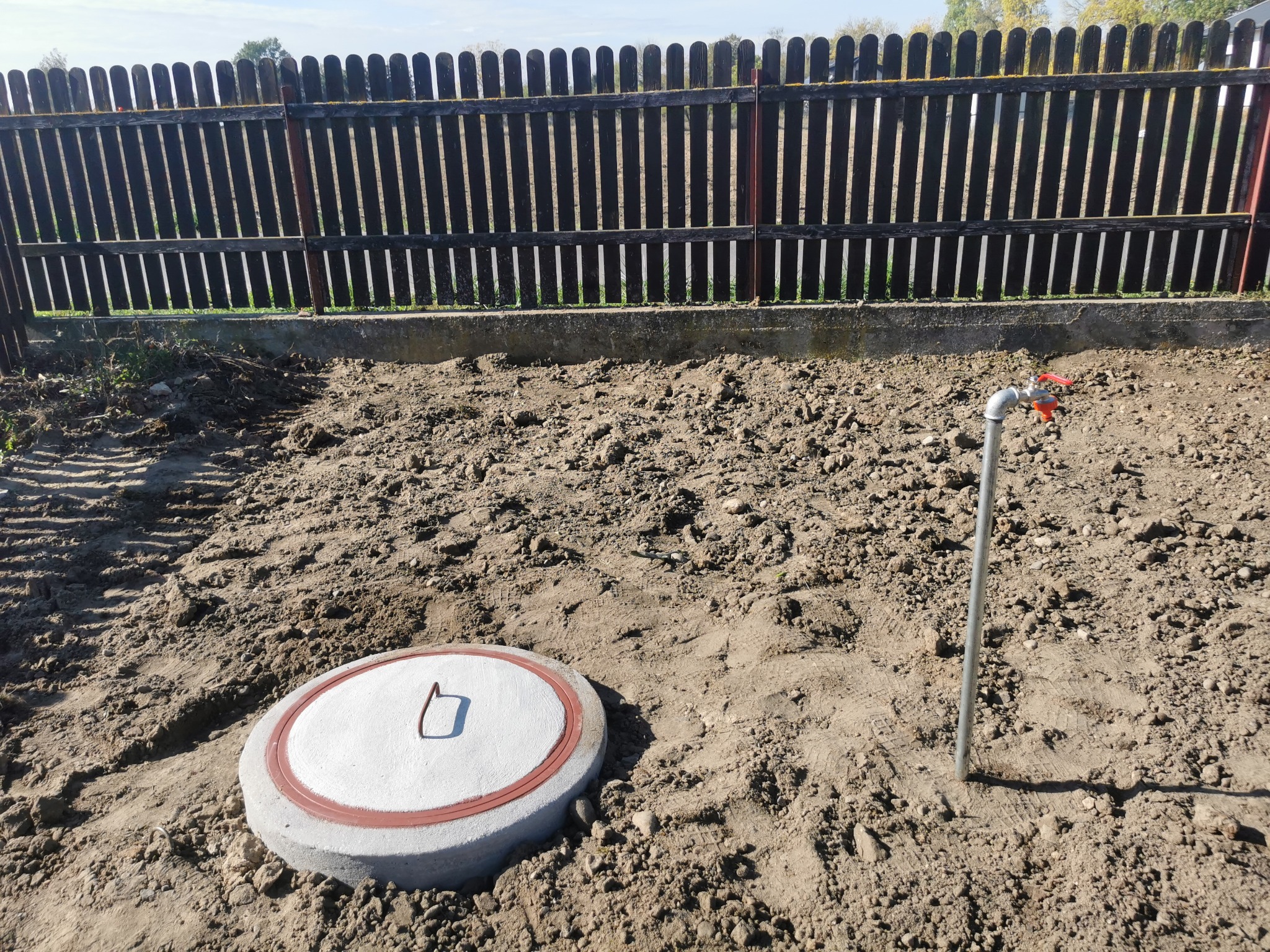 Betonowa pokrywa studni z metalowym uchwytem na piaszczystym gruncie, obok kran z czerwoną rączką i drewniany płot w tle.
