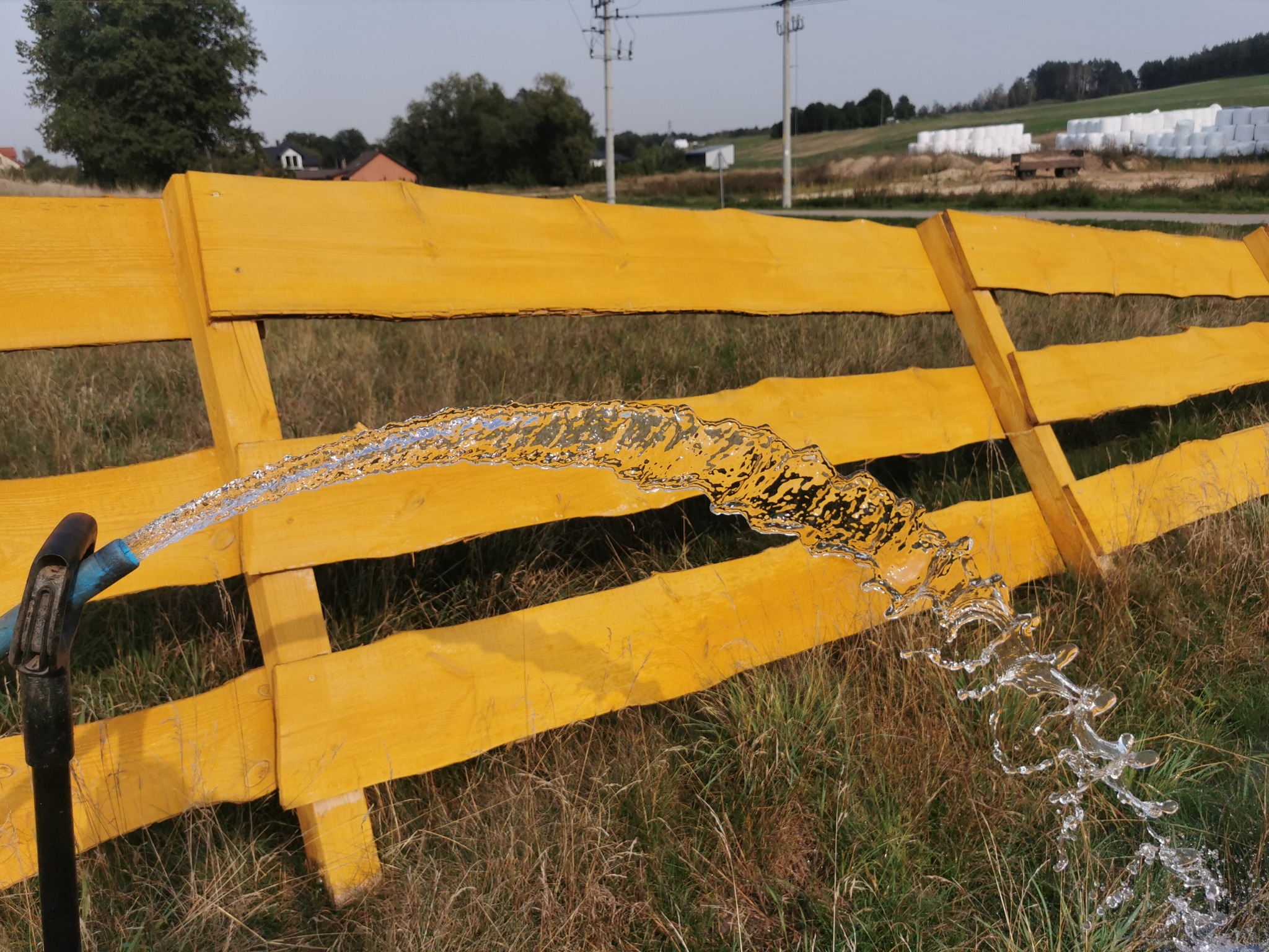 Strumień wody wypływający z niebieskiego węża, przelewający się przez żółte drewniane ogrodzenie na tle łąki i budynków.