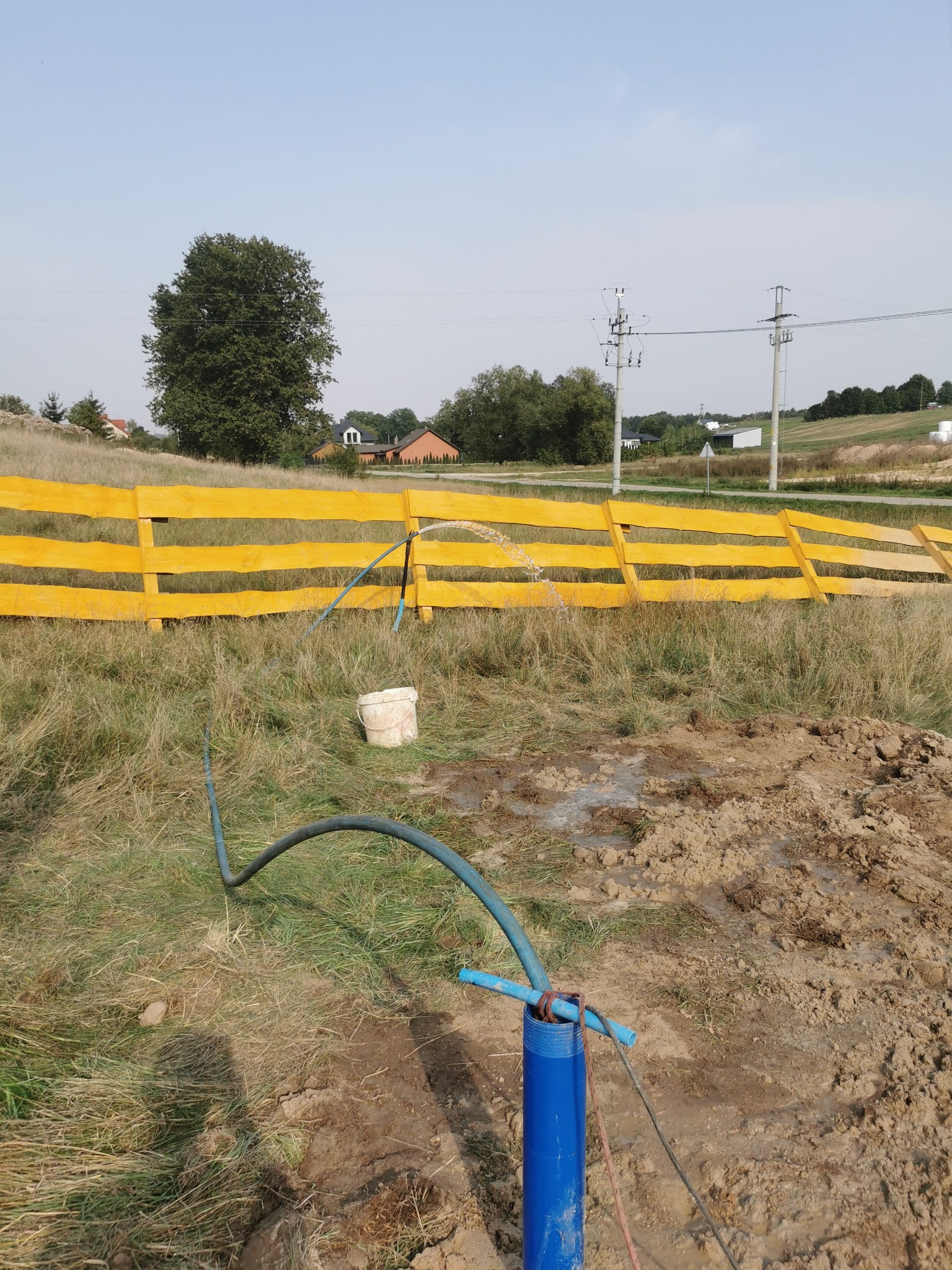 Woda tryskająca z niebieskiej rury wystającej z ziemi, z żółtym ogrodzeniem w tle, podczas wiercenia studni na zewnątrz.