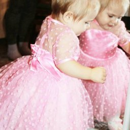 Ta/Na Fashion Sp. z o.o. - Dziewczynka w różowej tiulowej sukience z kokardą na plecach, odbijająca się w lustrze, z wzorem w małe serduszka na materiale.