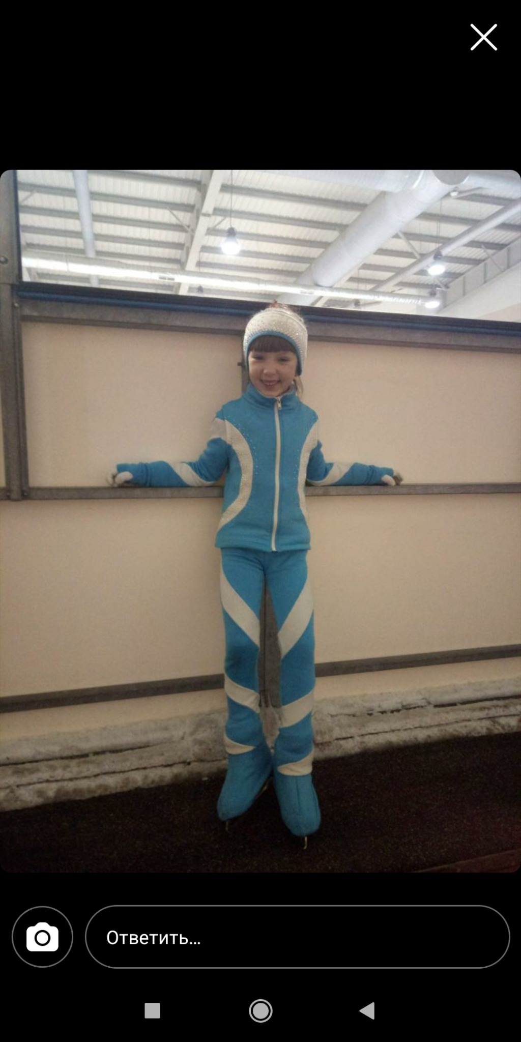 Uśmiechnięta dziewczynka w błękitnym, sportowym stroju z białymi wstawkami i dopasowanej czapce, stojąca na łyżwach przy białej ścianie lodowiska.