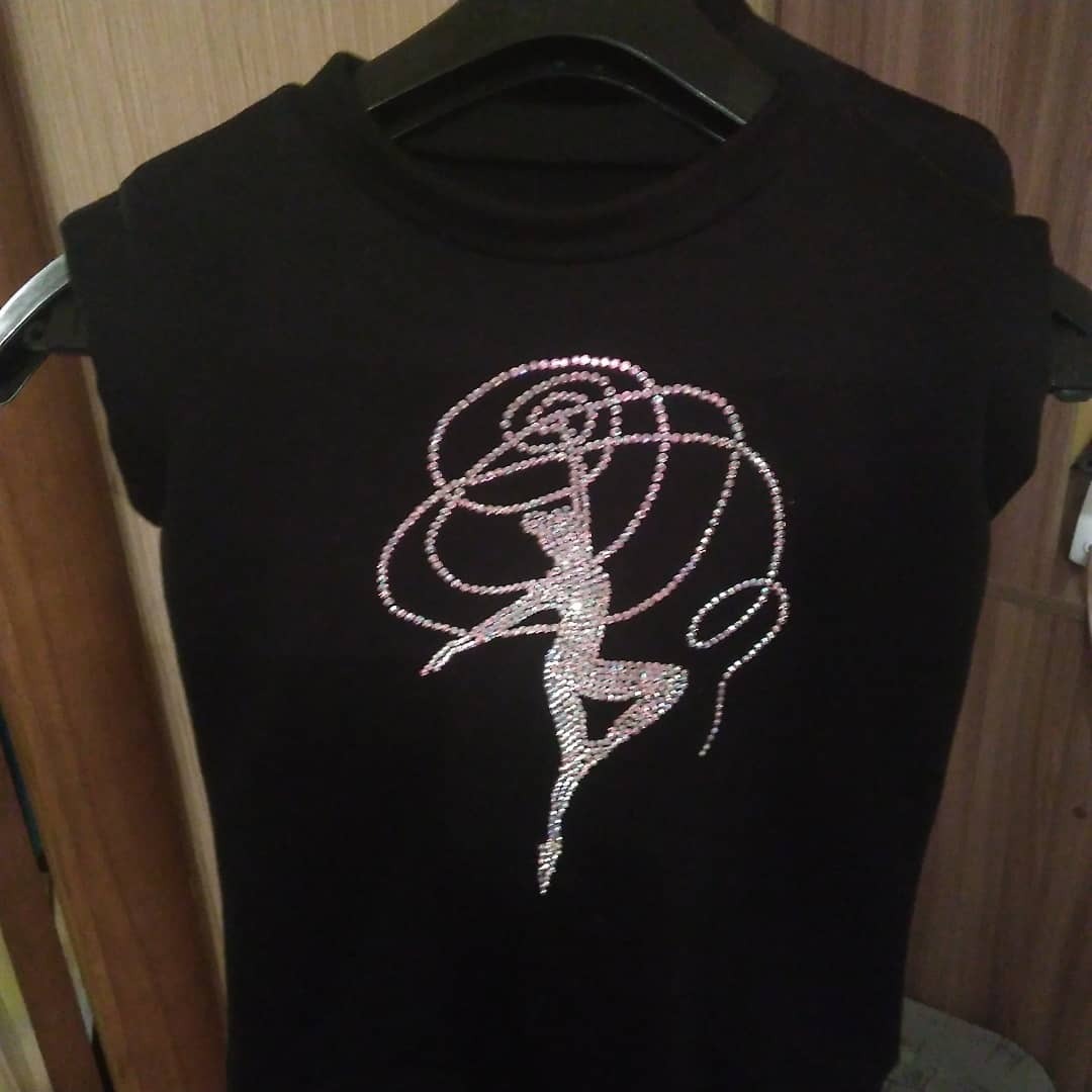 Czarna koszulka z aplikacją baletnicy i wstążki wykonaną z błyszczących, srebrnych i różowych koralików, wisząca na wieszaku.