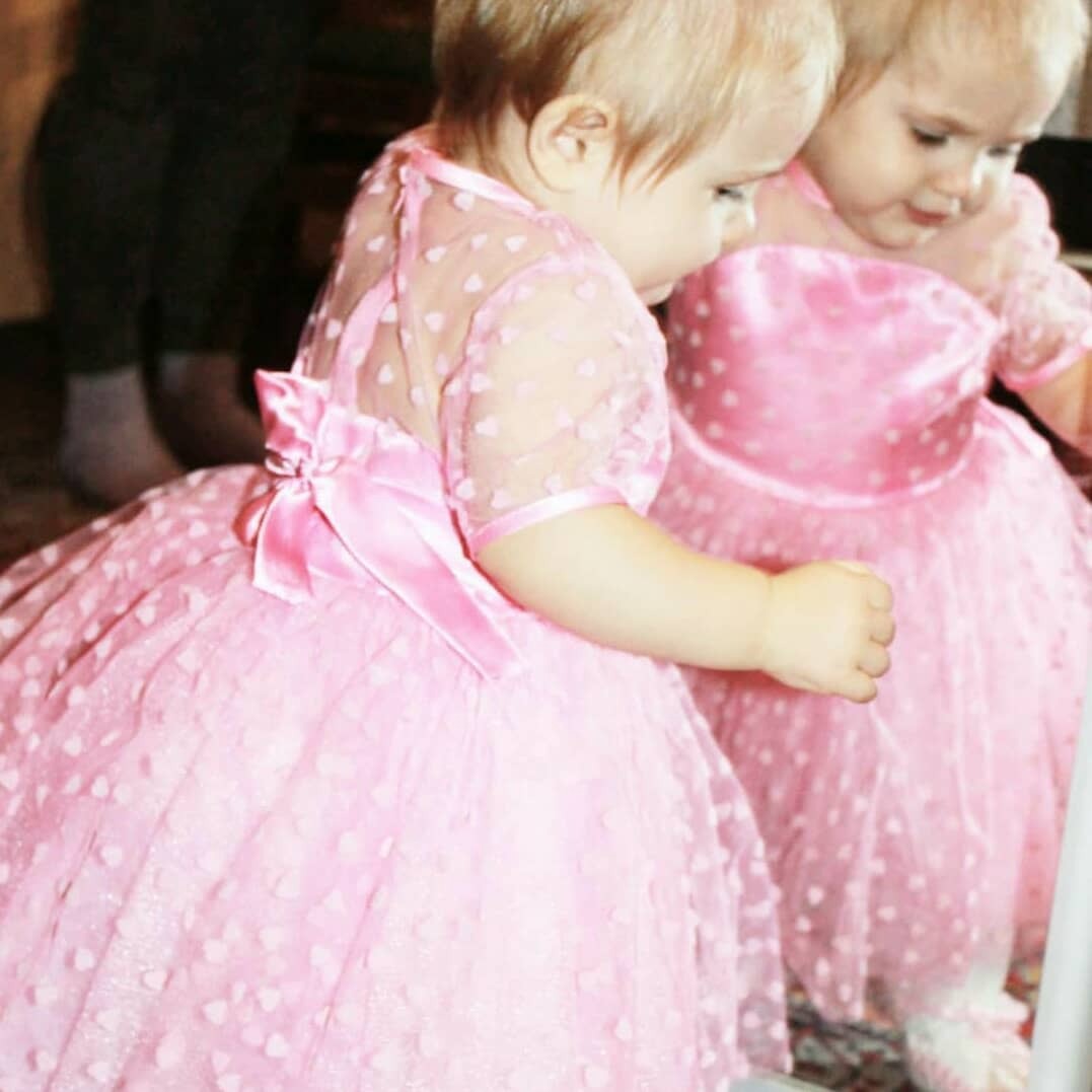 Dziewczynka w różowej tiulowej sukience z kokardą na plecach, odbijająca się w lustrze, z wzorem w małe serduszka na materiale.