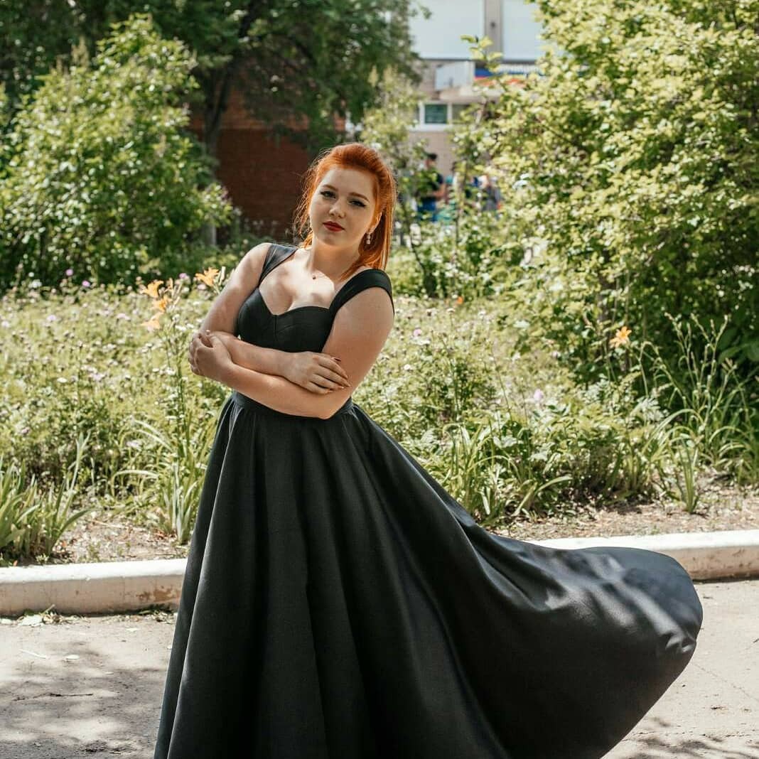 Ruda kobieta w czarnej, balowej sukni z rozłożystym dołem, pozująca na tle zieleni w plenerze.