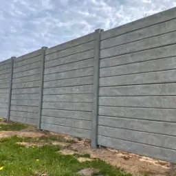 Szara, betonowa konstrukcja ogrodzeniowa z poziomymi panelami, widok z niskiej perspektywy w Ropczycach, z zielenią w tle.