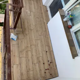 Układanie drewnopodobnych płytek na balkonie, widoczne krzyżyki dystansowe i poziomujące, przy oknie folia ochronna.