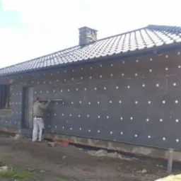 Pracownik mierzy poziom na elewacji domu jednorodzinnego z zamontowanymi płytami izolacyjnymi, widoczne białe kołki mocujące, dach pokryty ciemną dachówką ceramiczną, teren wokół domu w trakcie...