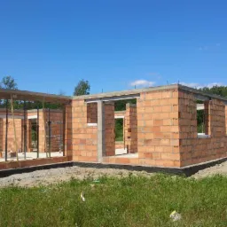 Wznoszenie ścian parterowego domu z czerwonej cegły, widoczne zbrojenie nad otworami okiennymi, na fundamencie zabezpieczonym czarną folią, w tle las i błękitne niebo.