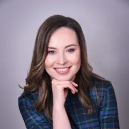 Portret uśmiechniętej kobiety w zielonej sukience w kratę, opierającej brodę na dłoni, na jasnoszarym tle.