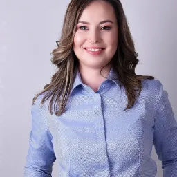 Portret uśmiechniętej kobiety w błękitnej koszuli i czarnych spodniach na jasnym tle. Kobieta ma brązowe włosy ułożone w fale.