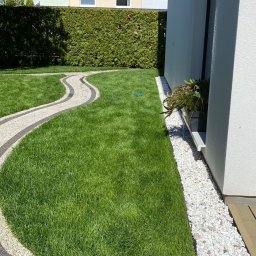 Paweł Iwanowski PafiNature - Świeżo przystrzyżony trawnik z krętą ścieżką z kostki brukowej, obrzeże z białych kamieni przy ścianie domu i żywopłot w tle.